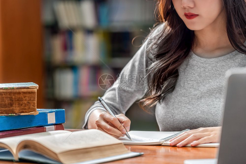 亚洲青年学生手工写功课和使用技术笔记本电脑在大学图书馆或具有各种籍和文具架背景上的各种群落大学图书馆或集体里写作业返回学校架子习图片