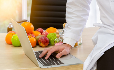 工作时穿白色实验室大衣的医生营养学家坐在木制桌子上用笔记本电脑键盘式计算机保健和饮食计划概念打膳咨询字吃苹果椅子图片