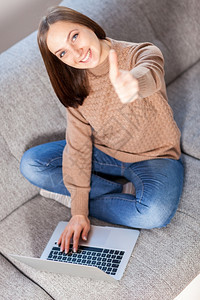 照片中笑着的caucasian女人在用电脑坐沙发上时做积极的拇指手势学生年轻的尽管图片