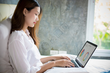 美丽亚洲妇女在家里工作时使用笔记本电脑她坐在床上科罗纳流行期间被隔离日冕的流行屋技术图片
