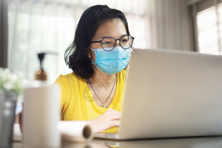 亚洲女设计师戴眼镜亚洲女设计师戴着眼镜在隔离期间与膝上型计算机举行在线视频会议孤立身穿黄色衬衫的青年女子则穿着蓝色外科面具在流行图片