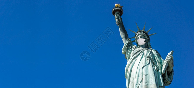 区著名的自由女神像戴外科面罩当时Covid19在全景或蓝色旗条背上爆发时佩戴外科面具纽约市美国corona流行建筑和与旅游概念约图片