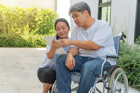 高级的坐在轮椅上妻子坐在旁边并拿着平板电脑病人因车祸而坐轮椅妻子在旁边让他花园漫步时放松一下不要让他看到与肩并和鼓励的观念引起亚图片