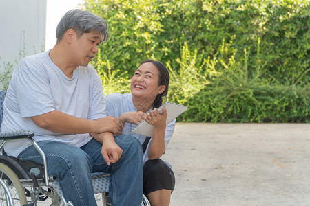 成人经过坐在轮椅上妻子坐在旁边并拿着平板电脑病人因车祸而坐轮椅妻子在旁边让他花园漫步时放松一下不要让他看到与肩并和鼓励的观念专家图片