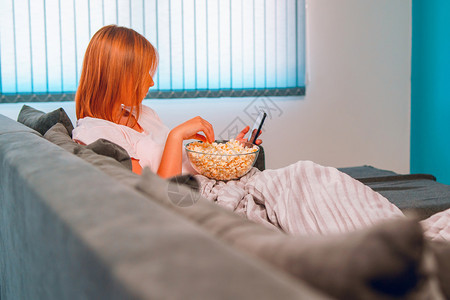 房间坐在家里睡沙发床上的年轻妇女躺在一个爆米花碗里在她公寓独自享受使用移动智能手机拍摄自照片或发送打字信息被盖着毯子的爆米花碗覆图片