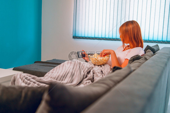 年轻的电话坐在家里睡沙发床上的年轻妇女躺在一个爆米花碗里在她公寓独自享受使用移动智能手机拍摄自照片或发送打字信息被盖着毯子的爆米图片