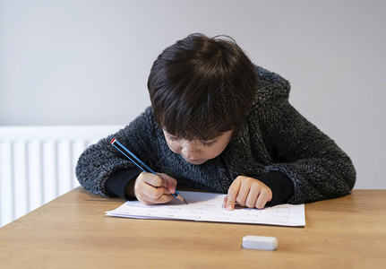 学校男孩坐在桌边做功课快乐儿童拿着铅笔写作一个男孩在白皮书小学和家庭教育概念上写英语字的画像男孩坐在桌上做功课男孩在白皮书上写英图片