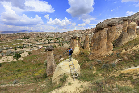 一个穿着蓝色T恤和背包的年轻男子站在卡帕多西亚的一个岩石上正看着他的手臂伸向山峡谷和蓝天空一名年轻男子站在一块岩石上看着卡帕多西图片
