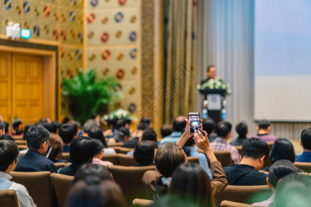 观众背面用手机照相发言人讲台和通过放映机在会议厅商业和教育概念的舞台上放映演示发言者讲台和演在会议室大厅商业和教育概念上电话亚洲图片