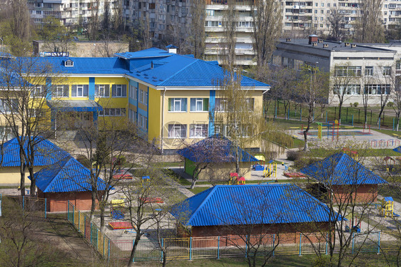 草树木幼儿园亮蓝色屋顶在灰城市高层建筑的背景下被春天阳光照亮幼儿园明的蓝色屋顶在灰城市高层建筑的背景下沙盒图片