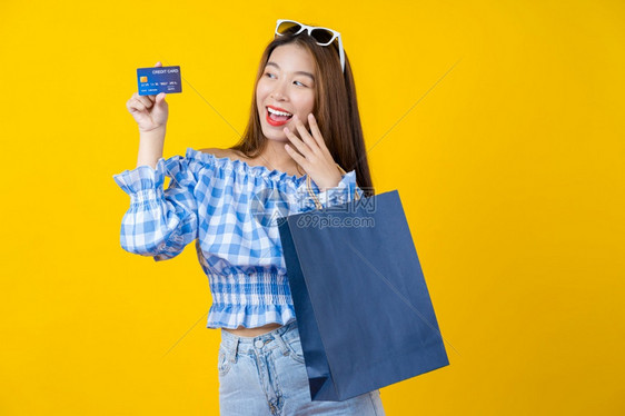 消费主义美丽快乐的亚洲微笑女使用信卡并携带一个购物大包装袋在孤立的黄色背景复制空间和工作室黑色周五销售上展示网购物同时提供黑星期图片