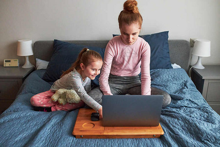 在家工作新冠流动在COVID19隔离期间在家庭笔记本电脑上远程用她的膝型电脑做学校测试作业的青少年女孩与坐在计算机前床上的小姐妹图片