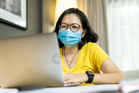 屋日本人亚洲女在家中隔离时使用膝上型计算机戴眼镜身穿黄色衬衫的青年女子戴着蓝色外科面具在流行Covid19科罗纳保健预防和护期间图片
