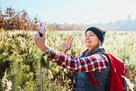 绿色假期照片女在视频电话中挥舞在山上度假旅行时致问候女背包徒步穿过高草地沿着的路走过渡与自然相亲的暑假图片