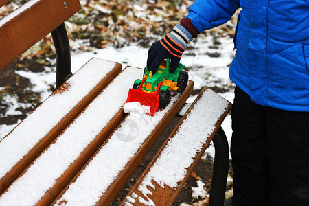 灰色的黑暗寒冷季节儿童游戏部分是小孩的画面站在被雪覆盖的木板凳旁孩子用玩具挖土机从板凳上扫清雪娱乐图片