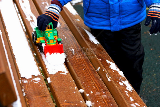 寒冷季节的儿童游戏部分是小孩的画面站在被雪覆盖的木板凳旁孩子用玩具挖土机从板凳上扫清雪多云的耙挖掘机图片