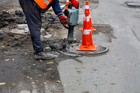 一名身穿反光橙色背心的道路工人用电动手提钻在被红色锥体包围的道路上沙井周围砸碎沥青复制空间一名道路工人用电动手提钻在道路上的下水图片