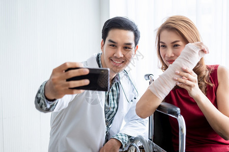 卫生年轻的夹板带着女病人戴手臂皮板的自拍坐在轮椅上使用智能手机在室内医院背景中坐着校对Portnoy图片