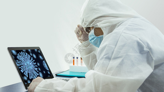 在职的关心带着蓝色面具和生物防护服的医在办公室里用笔记本电脑工作的侧视图他头靠在手上带着关切的态度看着屏幕上的白色背景的桌子上一图片
