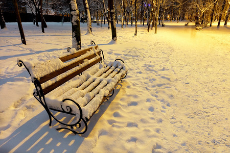 下雪的城市在公园冬夜在路灯暖光下雪地里空荡的棕色木凳上面有金属装饰栏杆季节图片