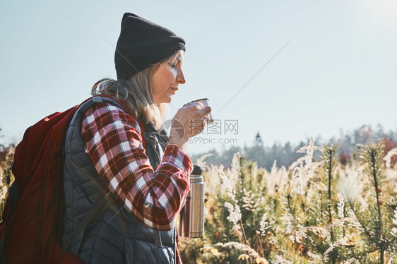 女子在暑假旅行中休息和放松喝着咖啡妇女站在足迹上看远女人背着包徒步穿越高草地在山区的道上漫步渡过与自然相亲的暑假高女热图片