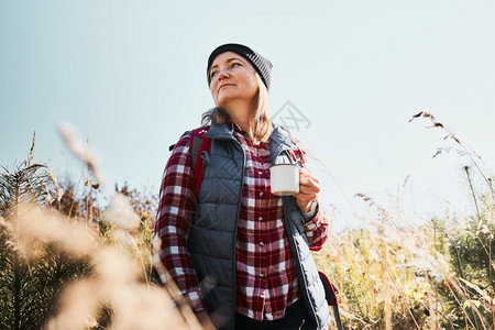 女子在暑假旅行中休息和放松喝着咖啡妇女站在足迹上看远女人背着包徒步穿越高草地在山区的道上漫步渡过与自然相亲的暑假春天秋热的图片