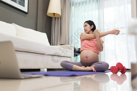 穿着粉红运动服装的亚洲有吸引力孕妇穿粉红色运动服在家中举起手来在家里做运动垫上瑜伽的年轻女而她在客厅怀孕时则运动垫上做瑜伽健康单图片