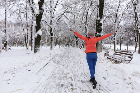 下雪的女孩身穿明亮珊瑚夹克和冬季蓝牛仔裤的年轻美女在雪覆盖的童话仙女公园大道上行走用木板凳子开着双臂张她的一名年轻美丽的少女在冬图片