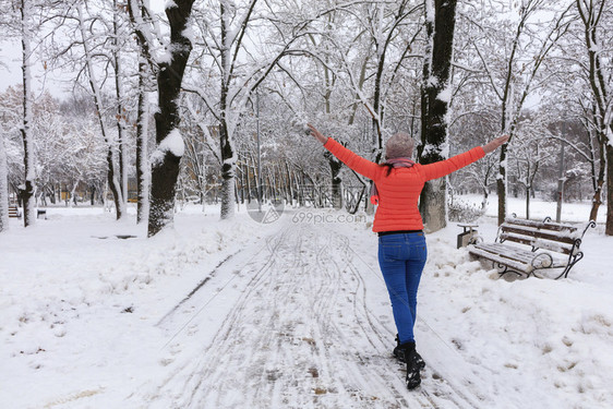 下雪的女孩身穿明亮珊瑚夹克和冬季蓝牛仔裤的年轻美女在雪覆盖的童话仙女公园大道上行走用木板凳子开着双臂张她的一名年轻美丽的少女在冬图片