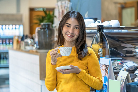 亚裔年轻女在现代咖啡店或工作间与咖啡机生活方式和休闲以及业余爱好和企家概念并列的咖啡店或合用空间一起喝咖啡电话衬衫闲暇背景图片