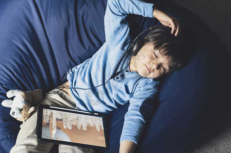 流行病阅读快乐男孩躺在豆袋上坐用数字平板关闭他的听音乐积极男孩留在家中休息隔离教育电子学习和自我分离期间在学校的网上家庭作业课后图片