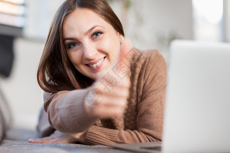 照片中微笑的女在躺沙发上与电脑聊天时使用正面的拇指手势内部的积极可爱图片