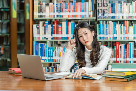 西班牙裔智力亚洲青年学生穿便衣思考如何做功课和在大学图书馆或群落使用技术笔记本电脑在书架上有各种籍和文具回学校读书时工作场所图片