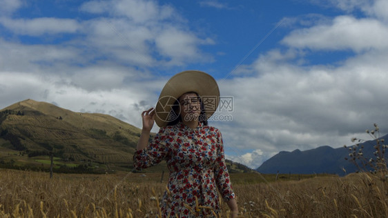 女士保持花的快乐年轻女子穿着花裙右手拿帽子独自走在麦田中间阴天从前面看到背景是蓝天和山脉在的阴手独自走在从前面看到的麦田中间图片