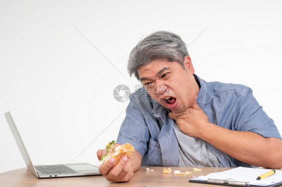 饮食汉堡包亚洲男子在办公桌上工作并吃汉堡在窒息食物后抱着颈部一个忙碌商人的概念无法保持工作左平衡也不能照顾健康只吃垃圾食品吧胖的图片