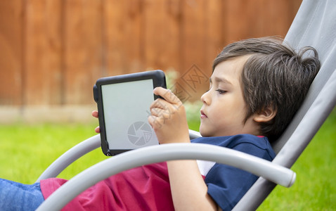 上学儿子平板玩游戏的肖像小孩一个在数字平板上看卡通时玩得开心的男孩子坐在摇椅上春天或夏的花园里放轻松户外图片
