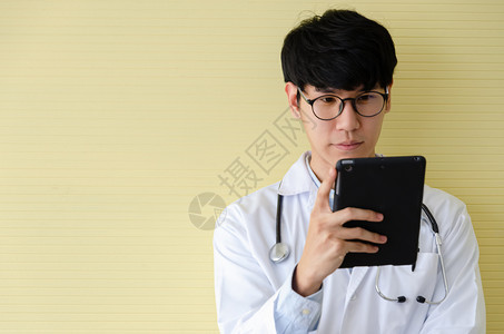 墙重点年轻男医生戴着眼镜站在黄色办公室里严肃地看着数字平板电脑的肖像健康检查的医疗保健概念例如为患者提供数字系统支持的测试结果成图片