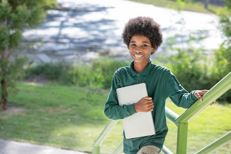 一个黑卷发的非洲男孩拥抱他的笔记本电脑微笑着对校舍下面的栏杆相机浓度联系摆姿势图片