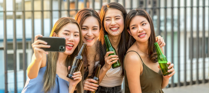 朋友们户外欢乐的亚洲女孩朋友团体在庆祝和跳舞时采取自拍在屋顶鸡夜节假日或一年度的周派对概念日落时与她的啤酒瓶一起庆祝和跳舞夜晚图片