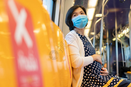 亚裔孕妇戴面罩脸具触摸肚子坐在天火车的黄色座位上同时前往具有社会距离的办公女商人室而大流行病为19岁或冠状黄色的卫生保健接触图片
