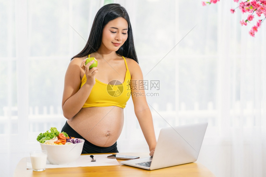 母亲腹部美丽亚洲怀孕妇辛勤工作吃饱并使用科技笔记本电脑蔬菜沙拉加绿苹果和早餐牛奶准备成为健康概念的母a亚洲孕妇辛苦工作吃饭和使用图片