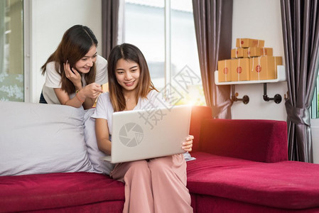 两名年轻的自由职业青年亚洲女企家在庭办公室私人工作手持膝上型计算机坐在沙发包装网上购物服务和向客户推销广告的网上邮包交付服务以及图片