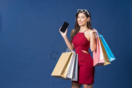 快乐的亚洲年轻女使用手机在蓝色背景无现金支付黑色星期五和消费主义概念下在线购物携带袋以手机买蓝色背景黑星期五和消费主义概念丰富多图片