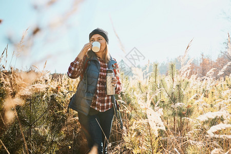 幸福步行健康在度假旅中享受阳光温暖的咖啡妇女站在高草地上望着远视举咖啡杯和热水瓶子的女人背着包徒步走在山里与自然相近的闲暇时间图片