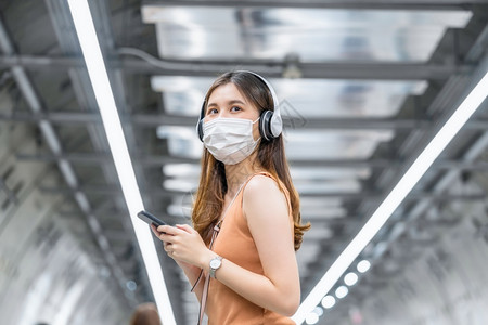 加冕在Covid19爆发感染和大流行病概念的都市旅行时身戴外科面具的亚洲年轻女乘客在地铁交接中通过智能移动电话监听音乐并在Cov图片