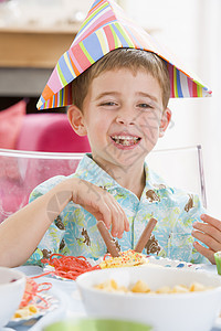 参加派对的年轻男孩坐在餐桌边 笑着食物生日孩子们男生微笑孩子男性桌子相机小男孩图片