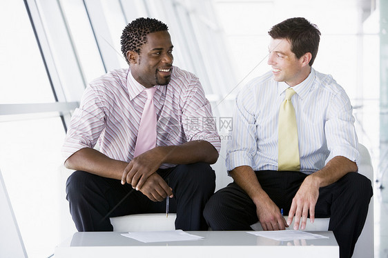 坐在办公室大厅前厅的两位商务人士在聊天和微笑年轻人同事两个人男性合作团队公司成人业务男人图片