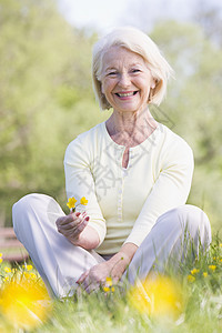 女人坐在户外微笑着笑着 拿着一朵 Buttcup花女性农村采摘场地毛茛女士公园花朵野花退休图片