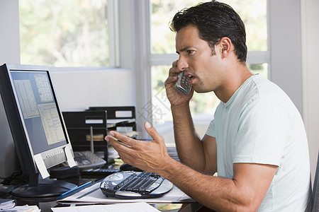 家庭办公室内使用计算机和眉毛打电话的男子图片