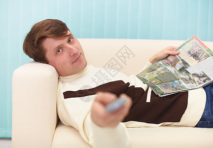 微笑的男人 躺在沙发上与杂志和电视遥控器一起图片